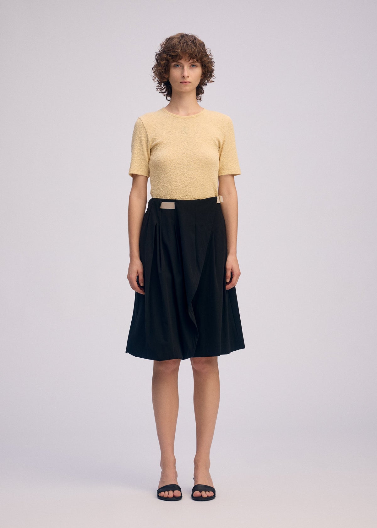 A person with short curly hair stands against a plain background, wearing the LOOPED short-sleeved light yellow textured top and black stretch knee-length draped skirt by ISSEY MIYAKE, paired with black open-toed sandals. view 5 of 8