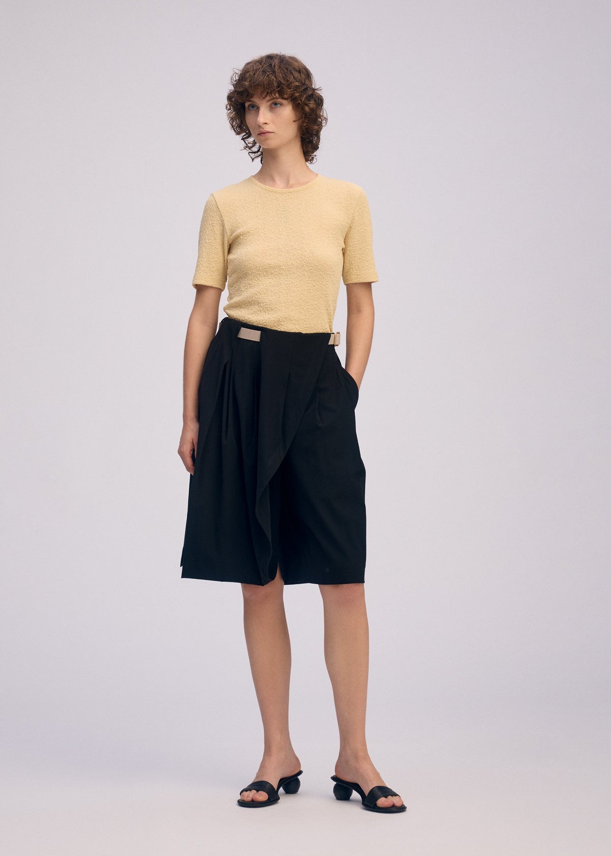 A person with short curly hair wears ISSEY MIYAKE's LOOPED: a light yellow short-sleeve top and black playful pleated shorts, paired with black open-toe sandals against a plain light background. view 6 of 8