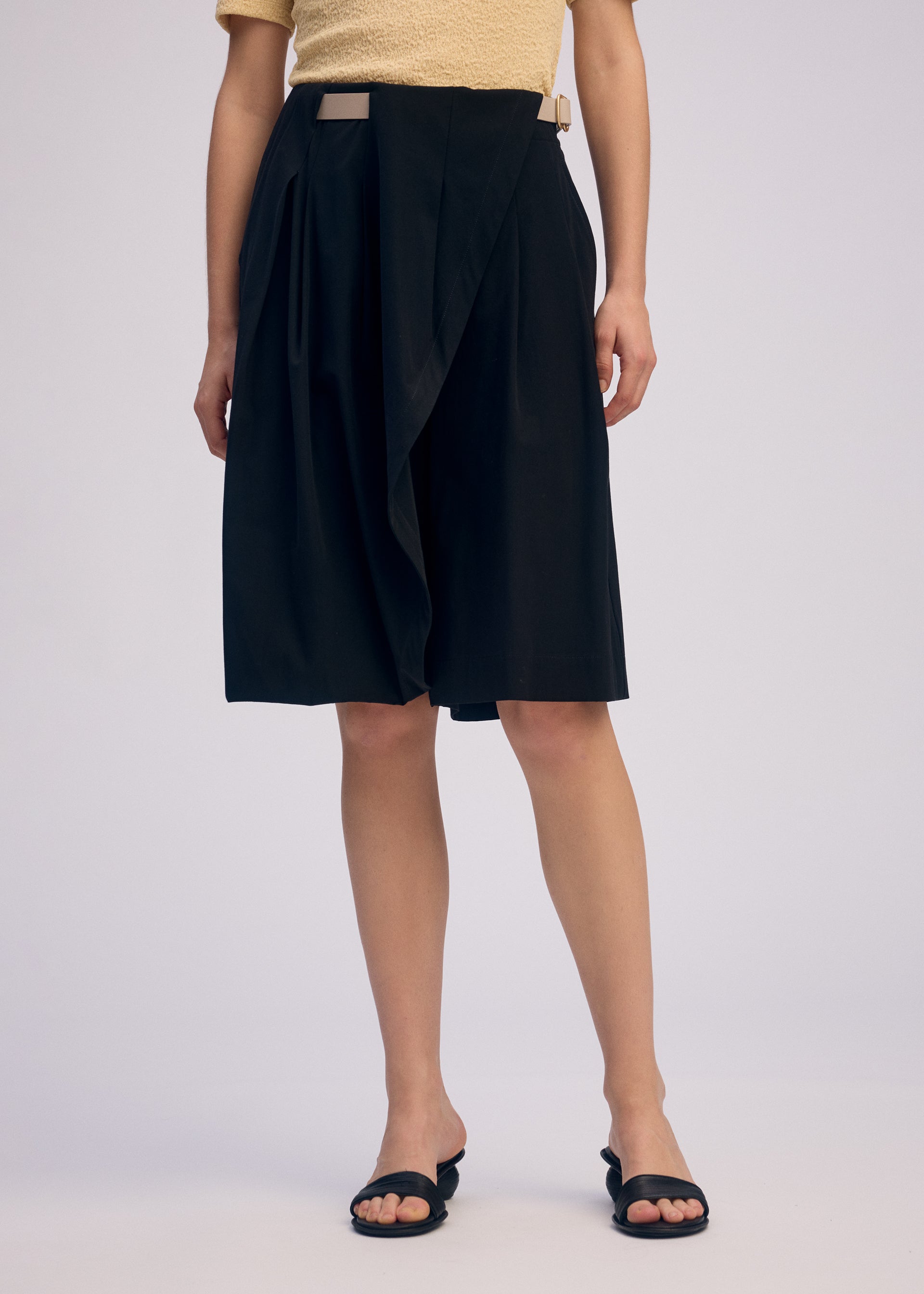 Zoomed view A person models the ISSEY MIYAKE LOOPED black knee-length skirt featuring a unique belt loop and wrap design, paired with a light yellow textured top and black open-toe sandals, standing before a plain light background. Their head is not visible.