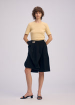 A person with short curly hair wears ISSEY MIYAKE's LOOPED: a short-sleeve beige textured top and black high-waisted knee-length skirt with unique belt loops, paired with black open-toe low-heel shoes, against a plain light background.