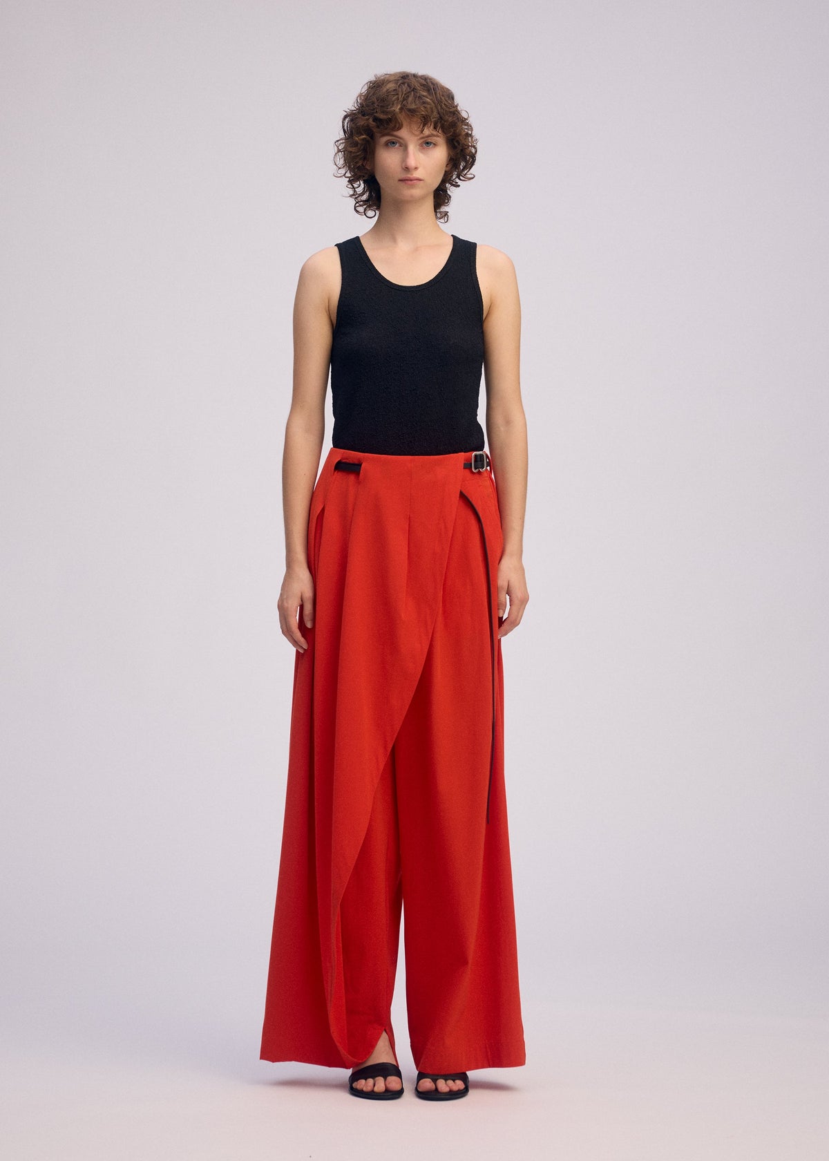 A person with short curly hair wears the ISSEY MIYAKE LOOPED wide-leg bright red pants with an asymmetric front flap, paired with a sleeveless black top and black sandals, standing against a plain light background. view 5 of 9