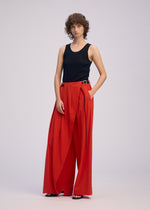 A person with short curly hair wears the ISSEY MIYAKE LOOPED sleeveless black top and high-waisted, belted wide-leg red pants, standing against a plain light background.