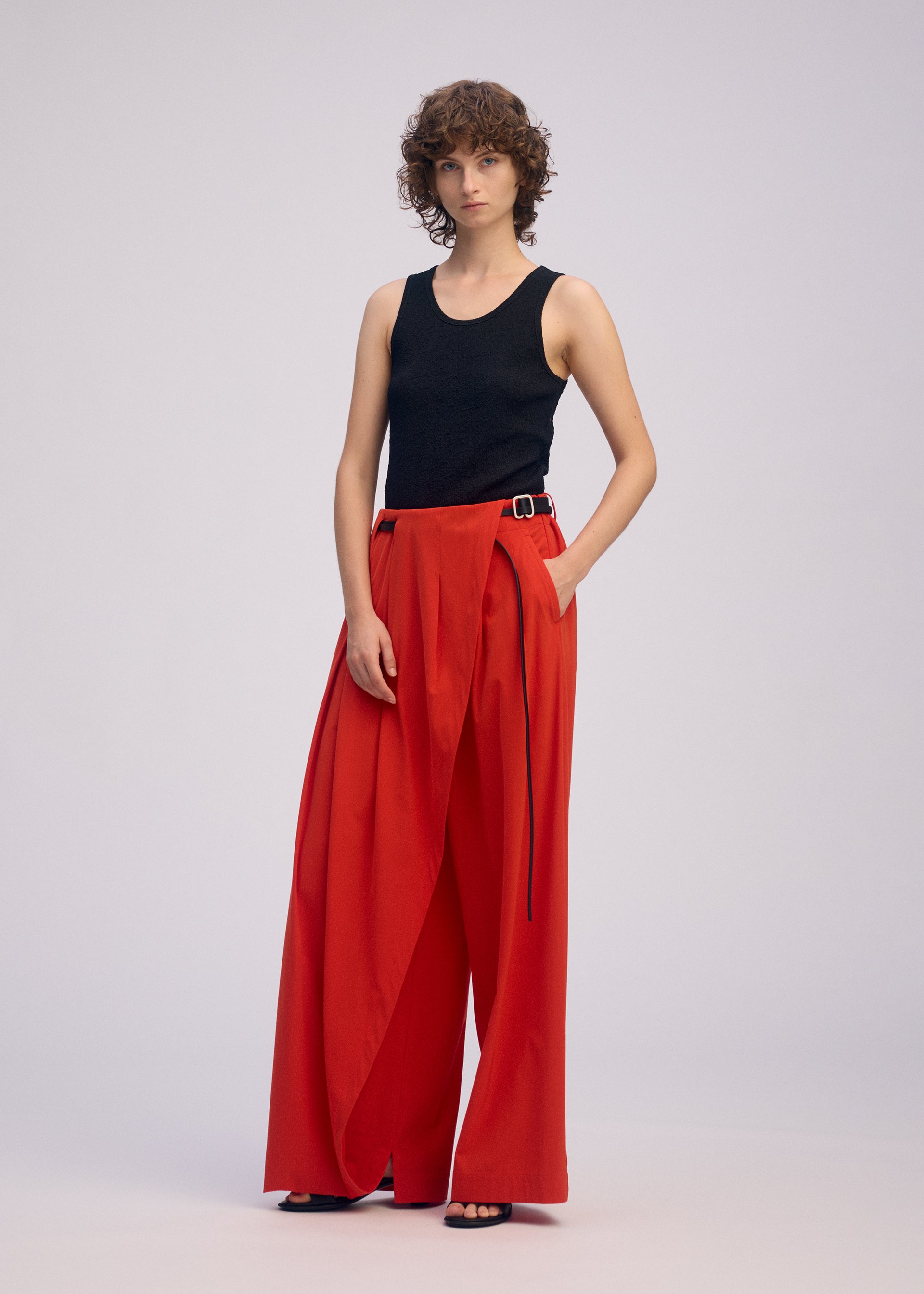 Zoomed view A person with short curly hair wears the ISSEY MIYAKE LOOPED sleeveless black top and high-waisted, belted wide-leg red pants, standing against a plain light background.
