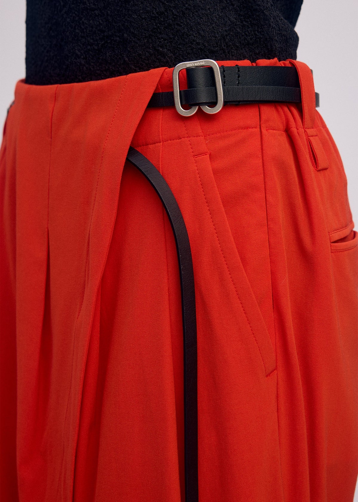 A close-up of a person wearing ISSEY MIYAKE's LOOPED high-waisted, loose-fitting red drapey stretch pants with pleats, styled with a black belt featuring a silver buckle and a textured black top tucked in. view 8 of 9