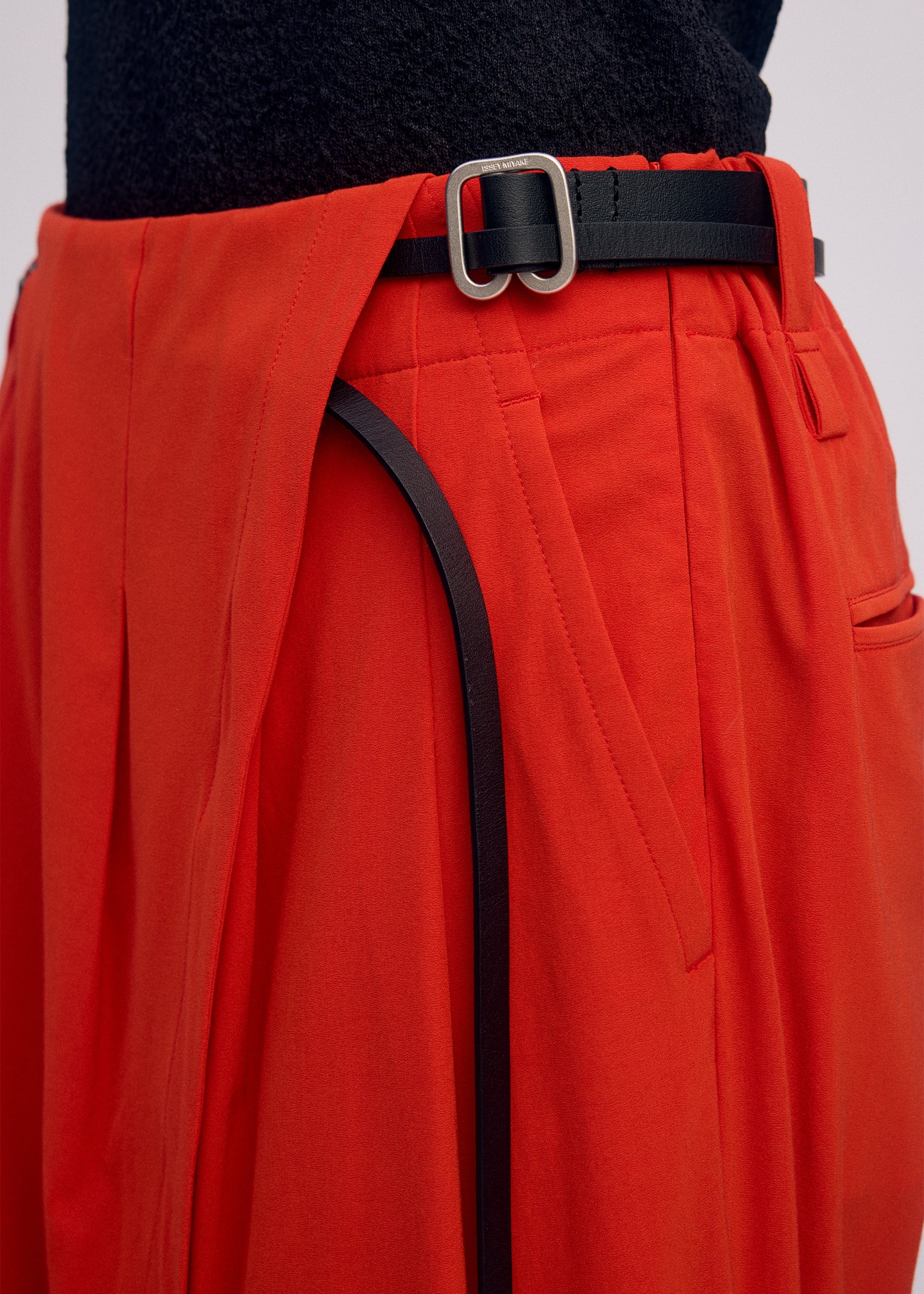 Zoomed view A close-up of a person wearing ISSEY MIYAKE's LOOPED high-waisted, loose-fitting red drapey stretch pants with pleats, styled with a black belt featuring a silver buckle and a textured black top tucked in.