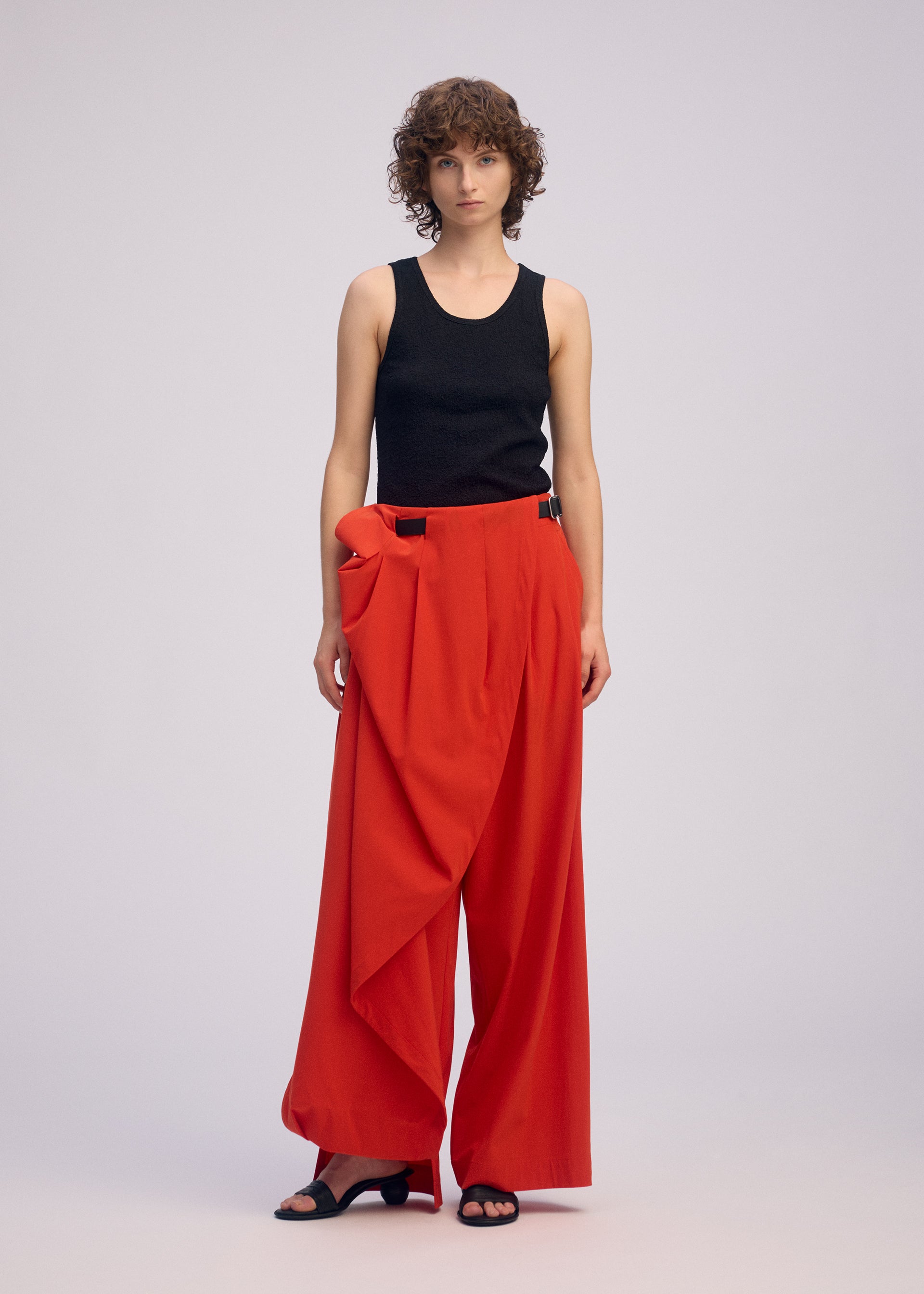 Zoomed view A person with short curly hair wears ISSEY MIYAKE's LOOPED red drapey stretch pants with unique belt loop detail, a black sleeveless top, and black open-toe shoes, standing against a plain light background.