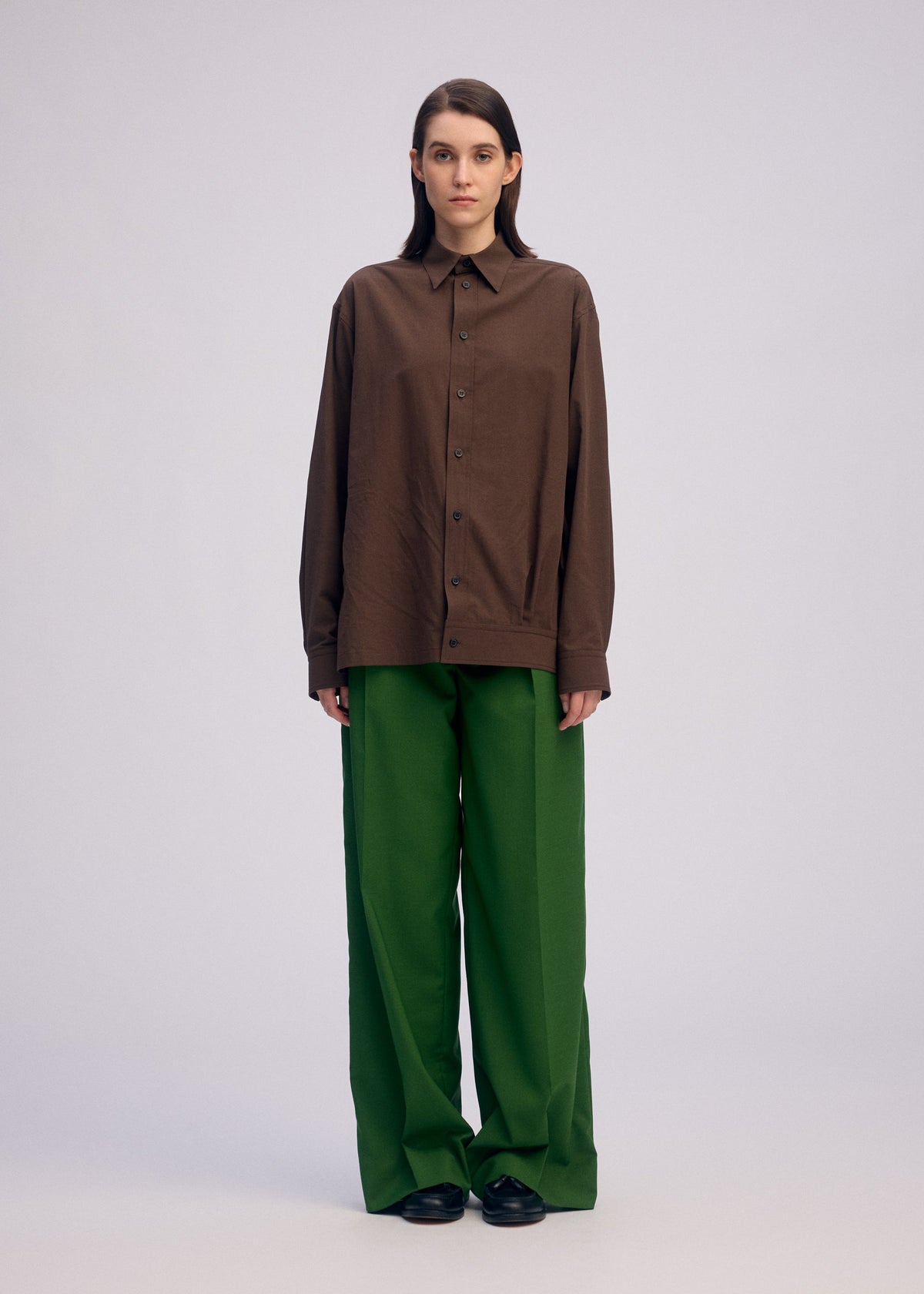A person stands against a plain light background, wearing ISSEY MIYAKE’s PALINDROME brown button-up shirt and wide reversible pants with black shoes. They have straight brown hair and a neutral expression. view 9 of 15