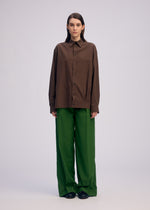 A person stands against a plain light background, wearing ISSEY MIYAKE’s PALINDROME brown button-up shirt and wide reversible pants with black shoes. They have straight brown hair and a neutral expression.