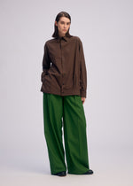 A person stands against a plain background wearing a loose brown button-up shirt, ISSEY MIYAKE PALINDROME wide-legged green reversible pants, and black shoes, with hands in pockets and a neutral expression.