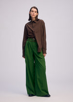 A person stands against a plain background in a loose brown button-up shirt and ISSEY MIYAKE PALINDROME wide-legged green reversible pants, paired with black shoes. Their hair is straight and shoulder-length.