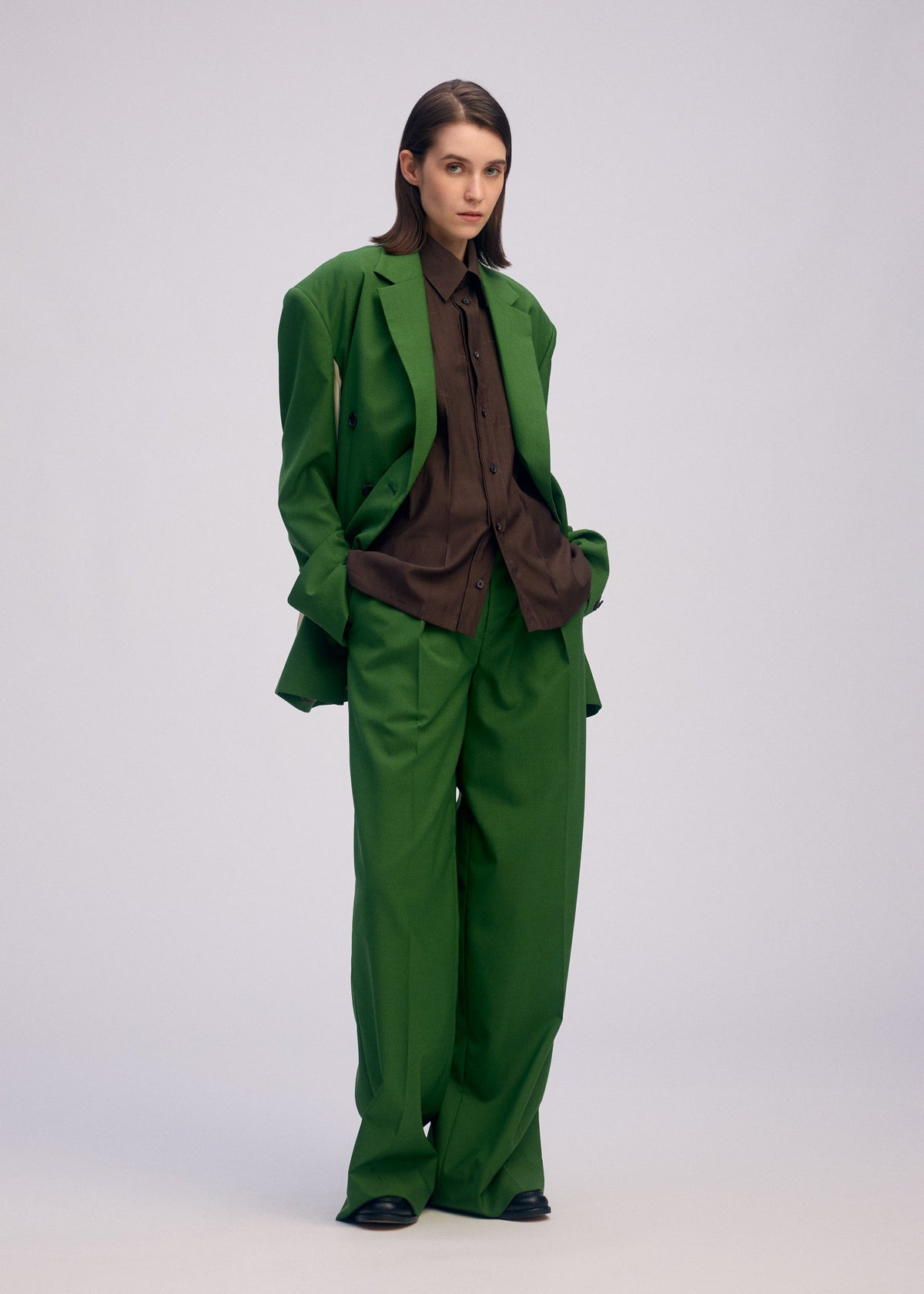 A person with straight brown hair stands against a plain background, hands in their pockets, wearing ISSEY MIYAKE's PALINDROME suit—loose-fitting with unique design and wide trousers—over a brown button-up shirt. view 12 of 15