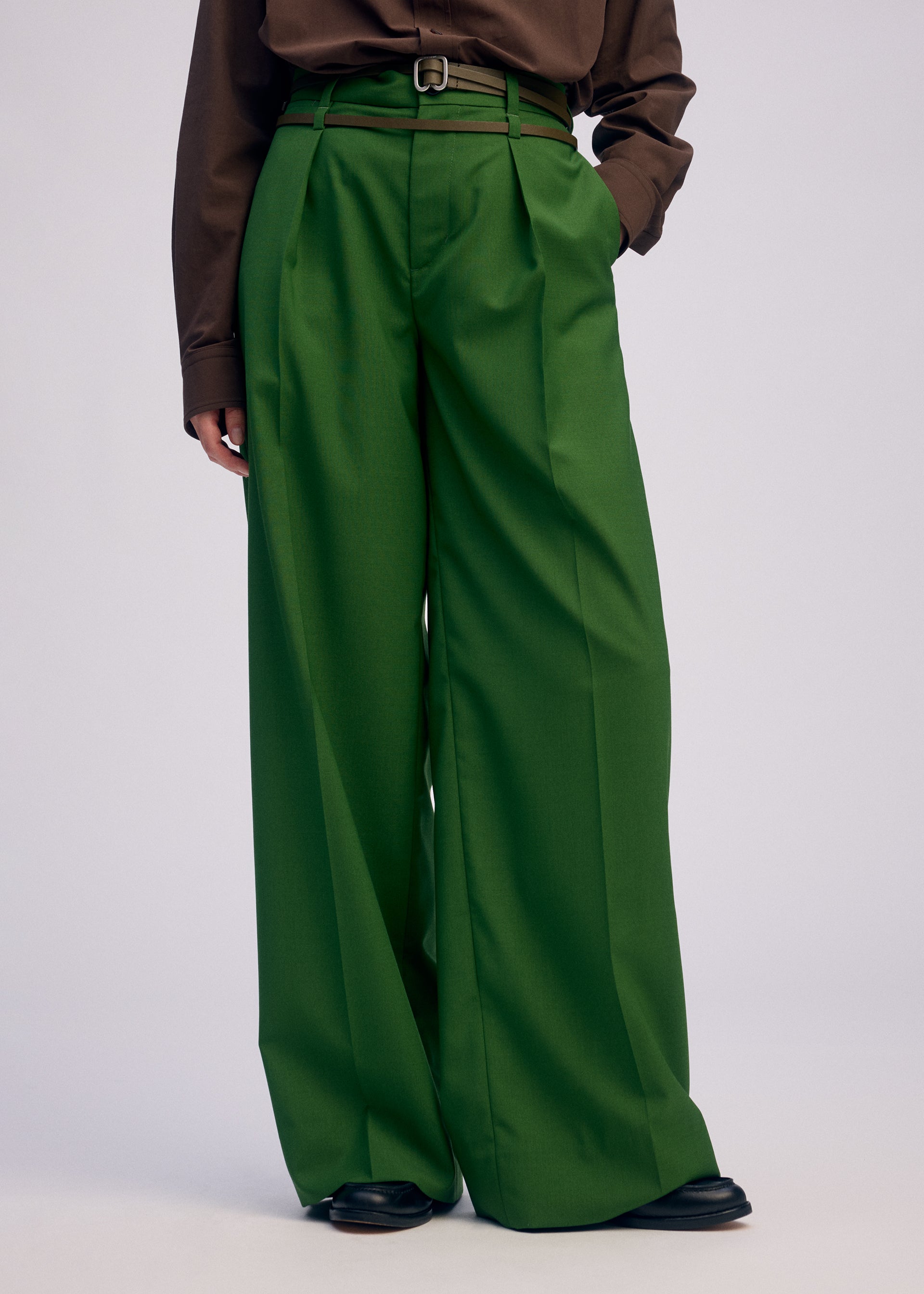 Zoomed view Wearing ISSEY MIYAKE’s PALINDROME high-waisted, wide-leg green trousers with a thin belt, brown long-sleeve shirt, and black shoes, a person stands against a plain background with one hand in their pocket.