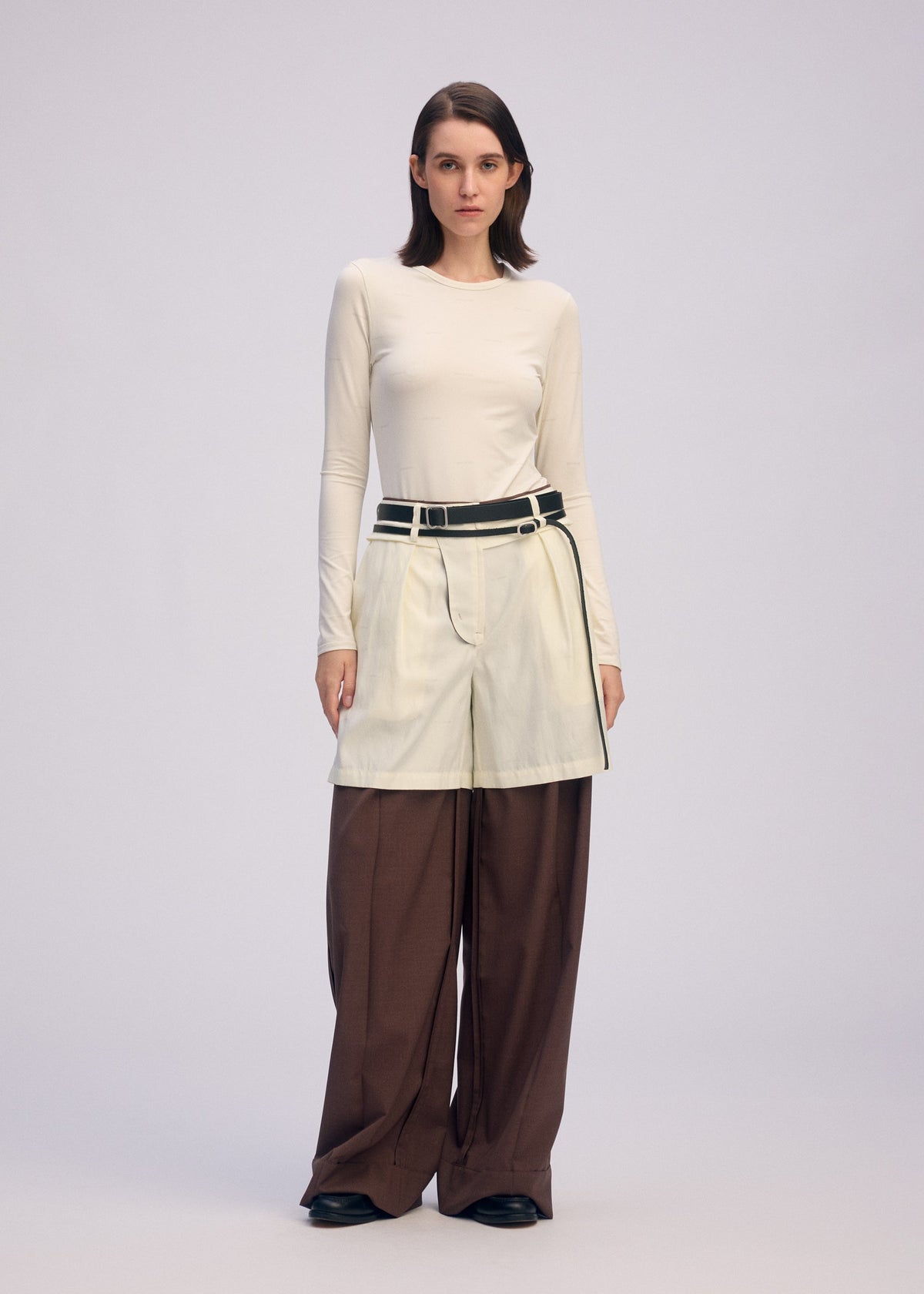 A woman stands facing forward in ISSEY MIYAKE’s PALINDROME: a fitted cream long-sleeve top, wide reversible pants, and a cream skirt with black belt layered over the trousers. The plain light background showcases the unique design. view 14 of 15