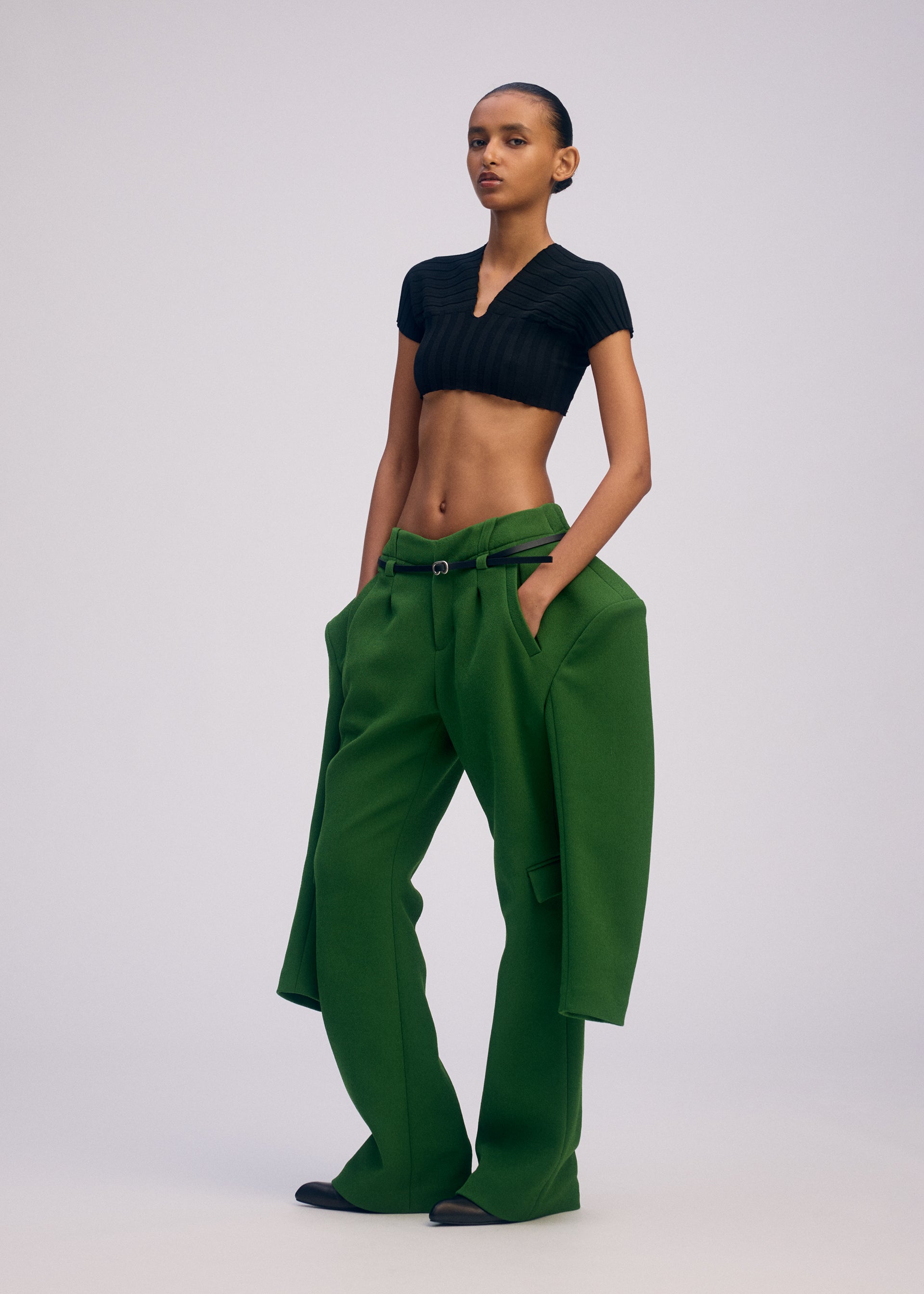 Zoomed view A person stands confidently in ISSEY MIYAKE’s ARMS oversized green triple-woven pants with a matching jacket at the hips and a fitted black crop top, the wide-legged suit striking against a plain, light-colored background.