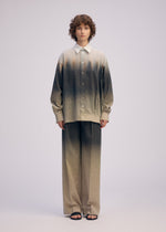 A person with short curly hair models the ISSEY MIYAKE PLUMAGE five-color gradient shirt and Kyoto artisan beige-and-dark pants, paired with black sandals, standing against a plain light background.