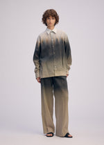 A person with short curly hair models the ISSEY MIYAKE PLUMAGE: a loose-fitting beige and gray gradient button-up shirt and Kyoto artisan five-color gradient pants, paired with black sandals, against a plain light background.