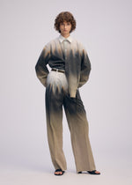 A person with curly brown hair wears the PLUMAGE shirt by ISSEY MIYAKE, featuring a five-color gradient pattern, paired with hand-painted neutral wide pants, black sandals, and a white belt against a plain light background.