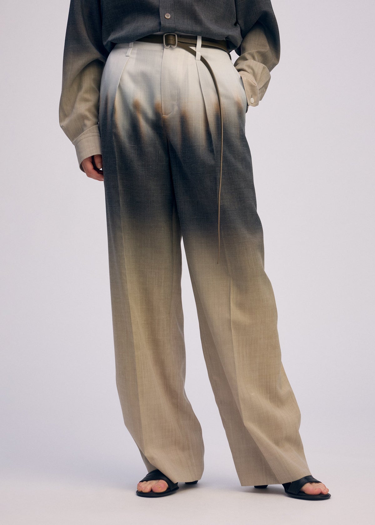 A person wears PLUMAGE wide pants and matching long-sleeve shirt by ISSEY MIYAKE, featuring a beige-to-black five-color gradient hand-painted by Kyoto artisans. The outfit is styled with a thin belt, black open-toe sandals, and a hand in the pocket. view 9 of 9