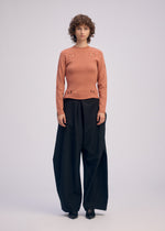 A person with short curly hair wears a salmon long-sleeved top with small cutouts and ISSEY MIYAKE FLUTTERING PANTS in black, standing against a plain light background.