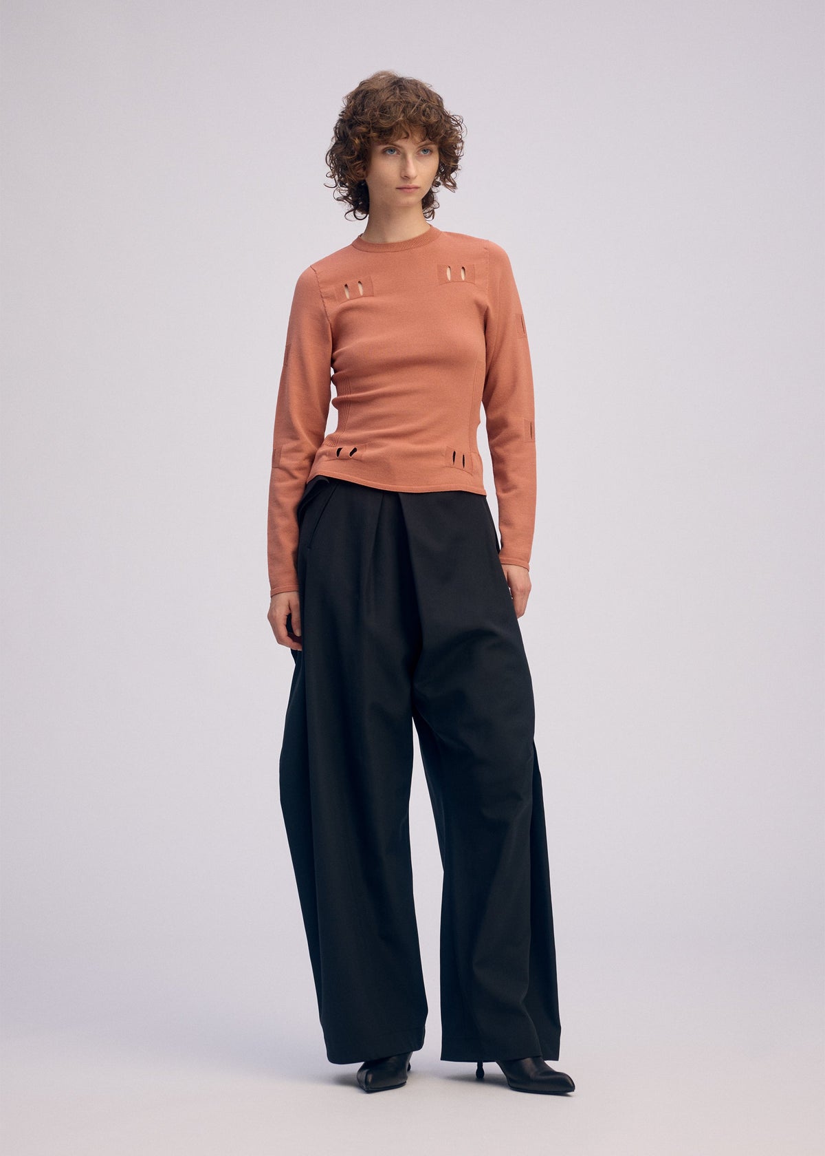 A person with short curly hair wears a fitted, long-sleeve peach top with cut-out details and ISSEY MIYAKE FLUTTERING PANTS in a wide-leg silhouette, paired with black heels, standing against a plain light background. view 7 of 9