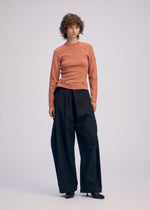 A person with short curly hair wears a fitted, long-sleeve peach top with cut-out details and ISSEY MIYAKE FLUTTERING PANTS in a wide-leg silhouette, paired with black heels, standing against a plain light background.