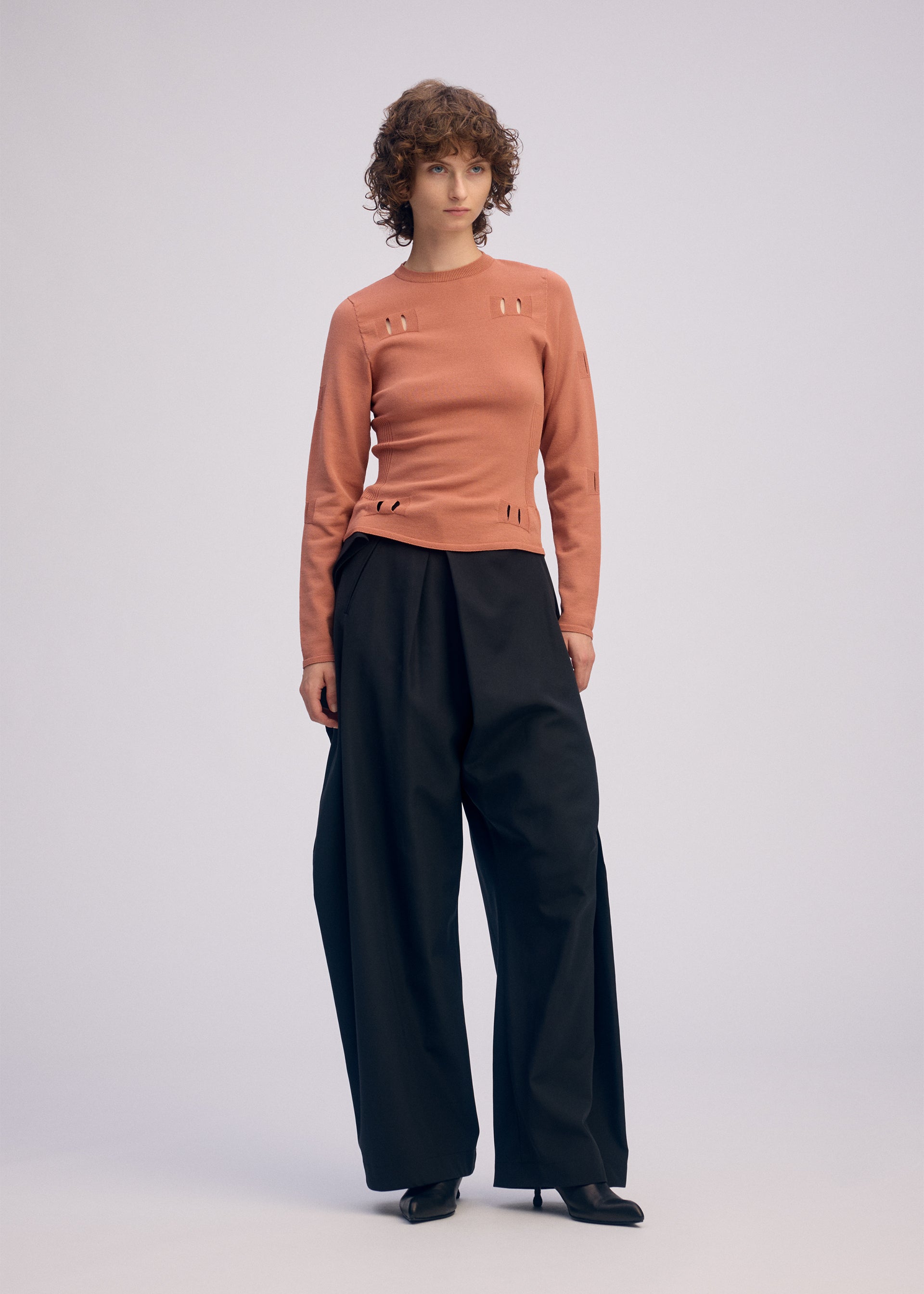 Zoomed view A person with short curly hair wears a fitted, long-sleeve peach top with cut-out details and ISSEY MIYAKE FLUTTERING PANTS in a wide-leg silhouette, paired with black heels, standing against a plain light background.