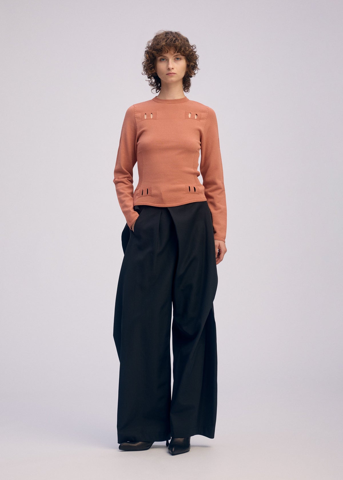 A person with short, curly hair and a neutral expression stands against a plain background, wearing a peach long-sleeved top with small cut-outs and ISSEY MIYAKE FLUTTERING PANTS in black. view 8 of 9