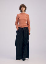 A person with short, curly hair and a neutral expression stands against a plain background, wearing a peach long-sleeved top with small cut-outs and ISSEY MIYAKE FLUTTERING PANTS in black.