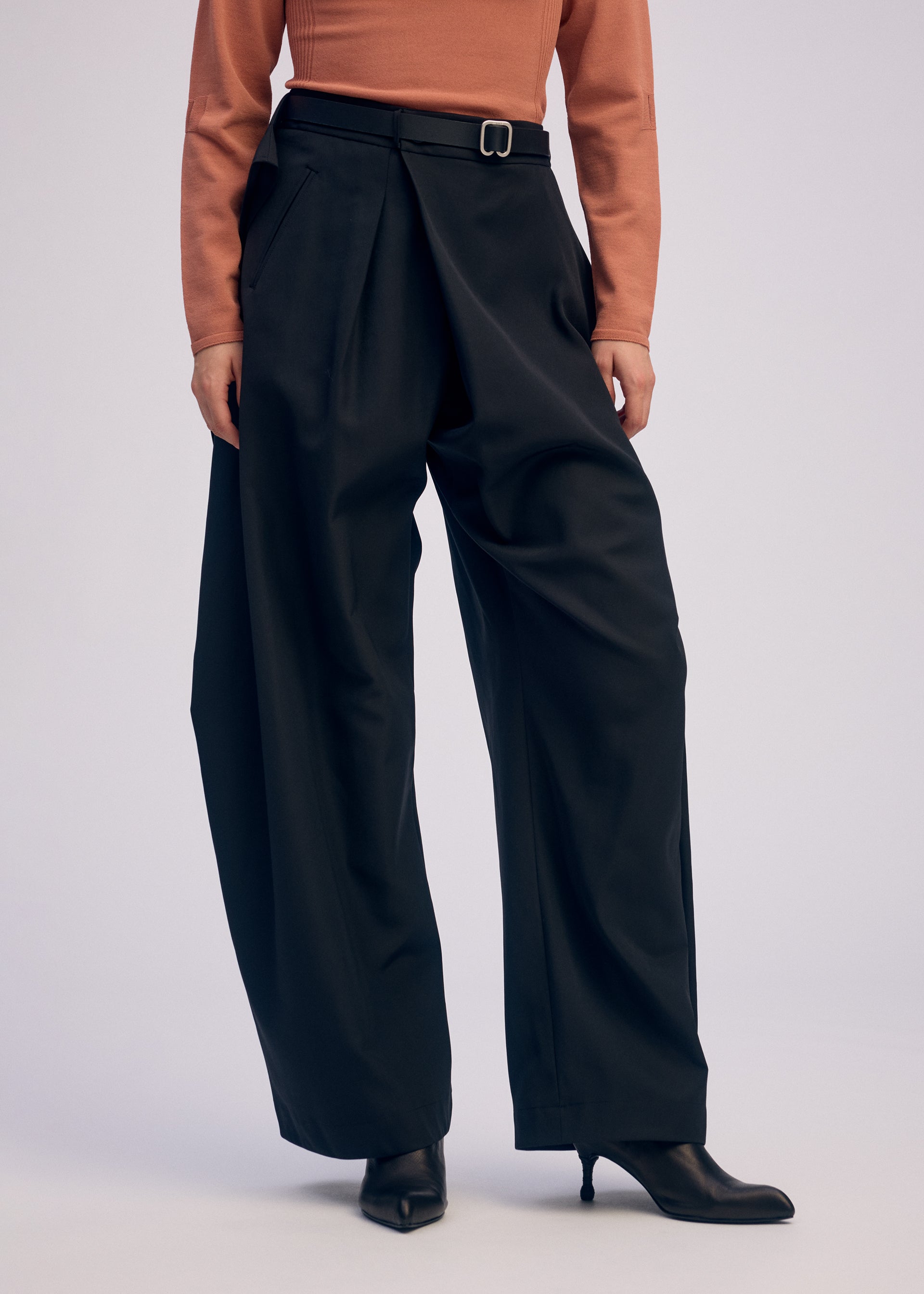 Zoomed view A person wears ISSEY MIYAKE's FLUTTERING PANTS—high-waisted, wide-leg, black, double-woven with pleats and a belt—styled with black heeled boots and a rust long-sleeved top. The cropped photo shows the matte textures complementing each other.