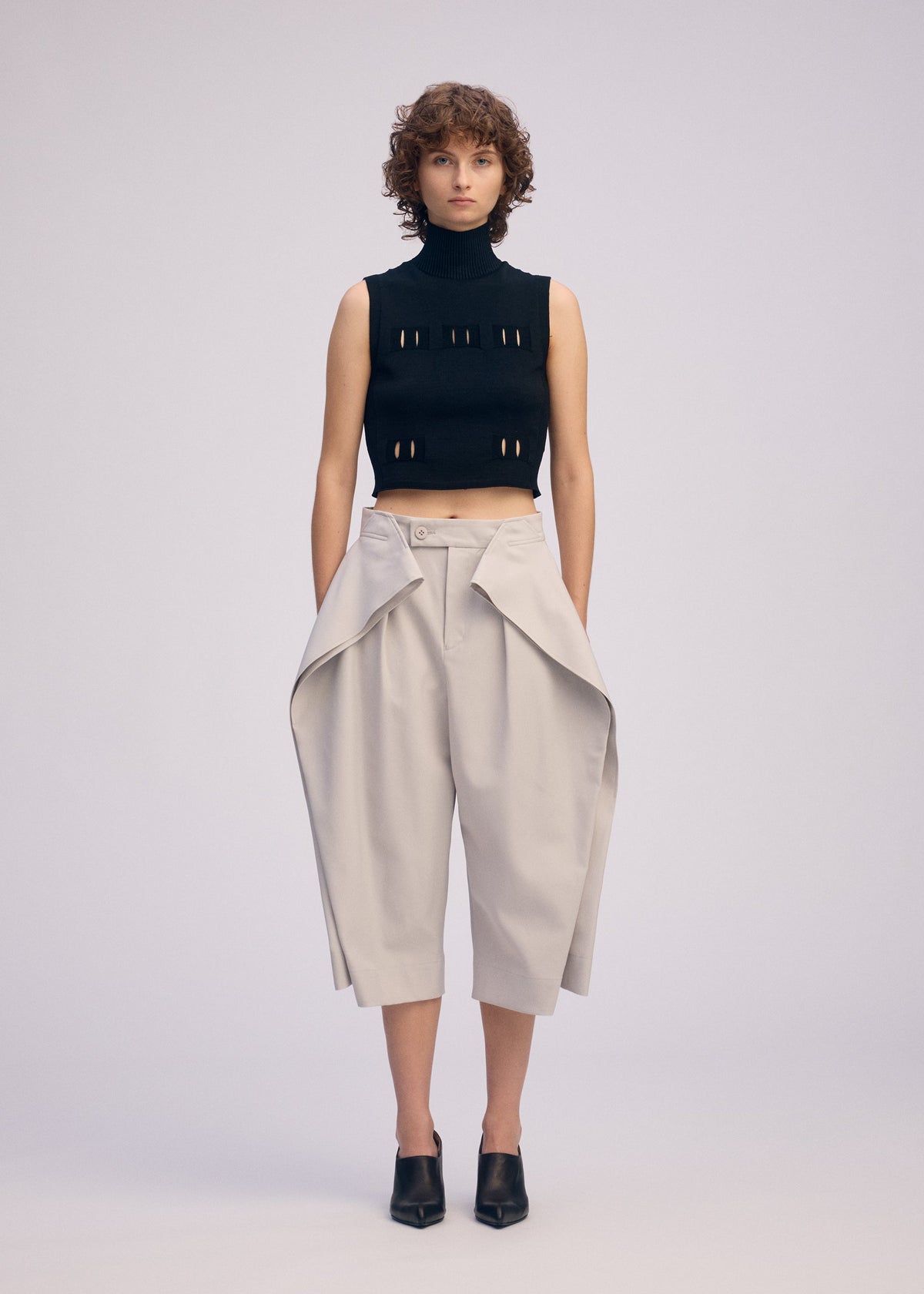 A person with short curly hair wears a sleeveless black turtleneck crop top and ISSEY MIYAKE FLUTTERING PANTS—light beige, high-waisted wide-leg pants with draped flaps—paired with black heeled mules, standing against a plain background. view 5 of 7