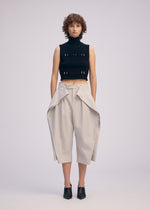 A person with short curly hair wears a sleeveless black turtleneck crop top and ISSEY MIYAKE FLUTTERING PANTS—light beige, high-waisted wide-leg pants with draped flaps—paired with black heeled mules, standing against a plain background.