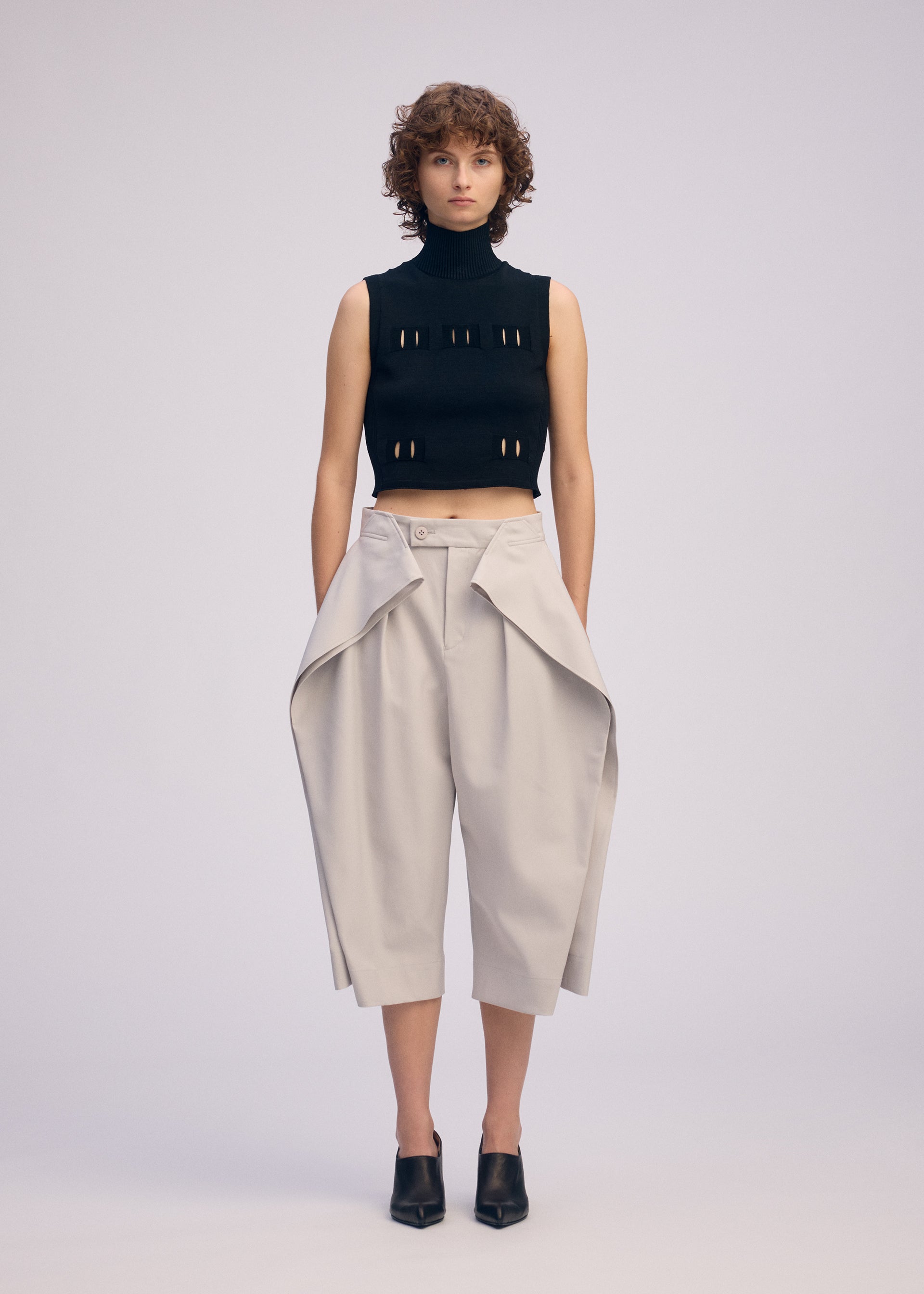 Zoomed view A person with short curly hair wears a sleeveless black turtleneck crop top and ISSEY MIYAKE FLUTTERING PANTS—light beige, high-waisted wide-leg pants with draped flaps—paired with black heeled mules, standing against a plain background.