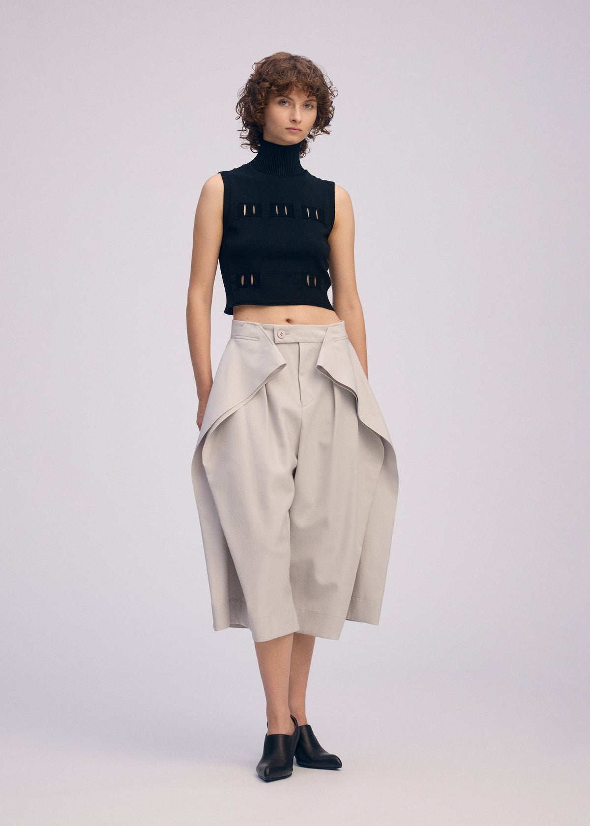 A person with short curly hair wears black mule shoes, a sleeveless turtleneck crop top with cutouts, and ISSEY MIYAKE FLUTTERING PANTS—wide high-waisted beige culottes with draped panels—against a plain light background. view 6 of 7