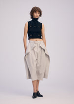 A person with short curly hair wears black mule shoes, a sleeveless turtleneck crop top with cutouts, and ISSEY MIYAKE FLUTTERING PANTS—wide high-waisted beige culottes with draped panels—against a plain light background.