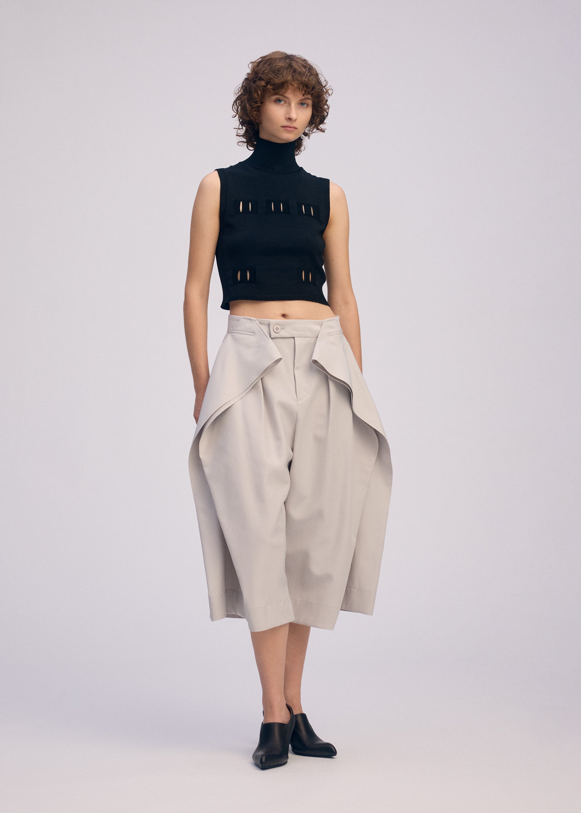 Zoomed view A person with short curly hair wears black mule shoes, a sleeveless turtleneck crop top with cutouts, and ISSEY MIYAKE FLUTTERING PANTS—wide high-waisted beige culottes with draped panels—against a plain light background.