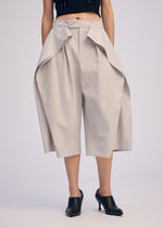 A person wears ISSEY MIYAKE FLUTTERING PANTS—wide, light beige culottes with dramatic draped panels in double-woven fabric—paired with black heeled mules, shown from waist to feet against a plain background.