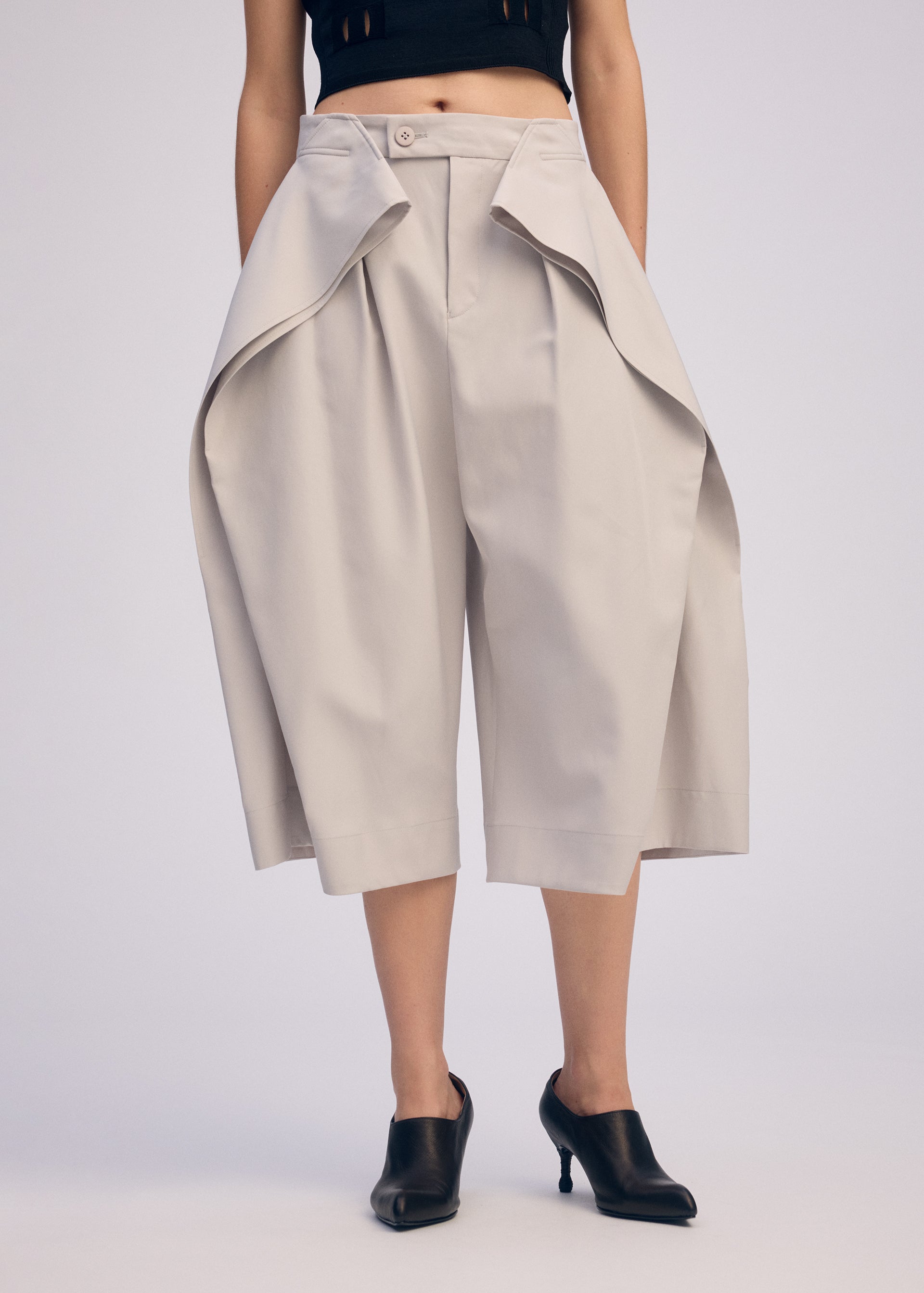 Zoomed view A person wears ISSEY MIYAKE FLUTTERING PANTS—wide, light beige culottes with dramatic draped panels in double-woven fabric—paired with black heeled mules, shown from waist to feet against a plain background.