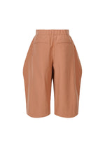ISSEY MIYAKE FLUTTERING PANTS: High-waisted, knee-length terracotta shorts with elastic waistband, front pleats, back welt pockets, and a subtle matte double-woven texture, shown against a white background.