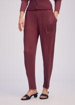 A person wears ISSEY MIYAKE SLEEK PLEATS maroon pleated tapered pants with a matching long-sleeve top, one hand in a pocket and black open-toe sandals. Image is cropped at the chest.