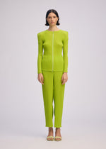 A person stands facing forward, wearing the ISSEY MIYAKE BOX SHOULDER PLEATS set—a bright lime green, long-sleeved, ribbed top with matching double-pleated pants—paired with open-toed sandals against a plain light background.