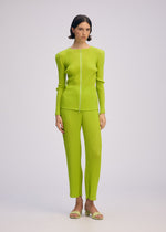 A person stands against a plain light background wearing the ISSEY MIYAKE BOX SHOULDER PLEATS set: a bright lime green ribbed, long-sleeve zip-up top with matching double-pleated pants, styled with open-toe sandals.