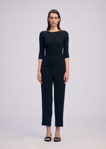 A woman faces forward in ISSEY MIYAKE’s RHOMBUS PLEATS: a fitted, textured black top with three-quarter sleeves and matching sleek black pants. She has straight shoulder-length brown hair, wears black sandals, and stands before a plain light gray background.