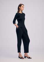 A woman stands in a studio wearing the ISSEY MIYAKE RHOMBUS PLEATS set—black, ribbed three-quarter sleeve top and loose pants with pockets—styled with black open-toe sandals, gazing to the side with a neutral expression.