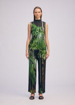 A woman with straight hair models ISSEY MIYAKE's URBAN JUNGLE PLEATS: a sleeveless, high-neck top and wide-leg pants with vivid palm tree print, paired with black open-toed sandals against a plain background.