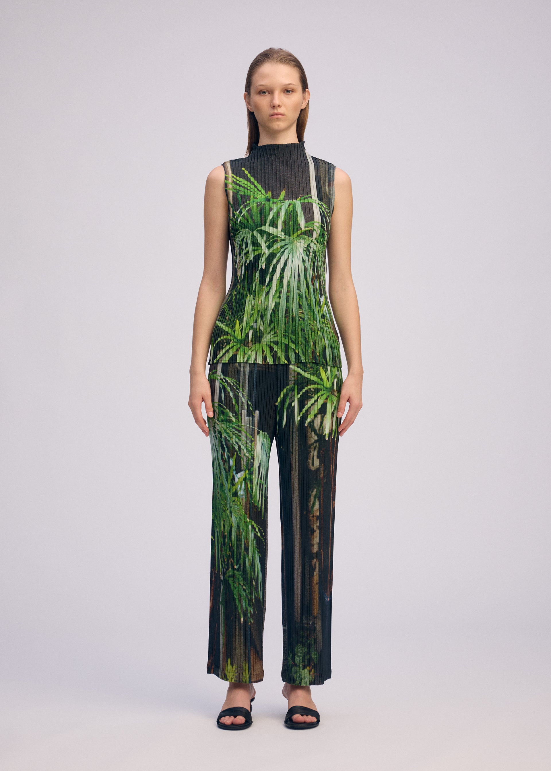 Zoomed view A woman with straight hair models ISSEY MIYAKE's URBAN JUNGLE PLEATS: a sleeveless, high-neck top and wide-leg pants with vivid palm tree print, paired with black open-toed sandals against a plain background.