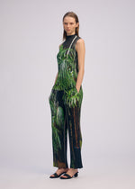A woman poses in URBAN JUNGLE PLEATS by ISSEY MIYAKE—a sleeveless, high-neck top and wide-leg palm print pants with hands in pockets, black sandals, set against a plain light background.