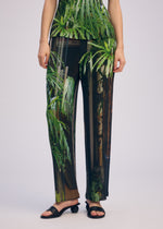 A person wears the ISSEY MIYAKE URBAN JUNGLE PLEATS set, showcasing a lush green tropical forest print, paired with black open-toe sandals. The image is cropped at the shoulders.