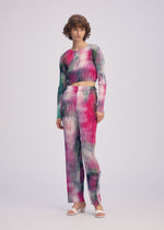 A person with short curly hair wears the OBSCURE PLEATS long-sleeve top and patterned pants from ISSEY MIYAKE, featuring a pink, green, and white striped tie-dye design. They stand against a light background in white sandals.