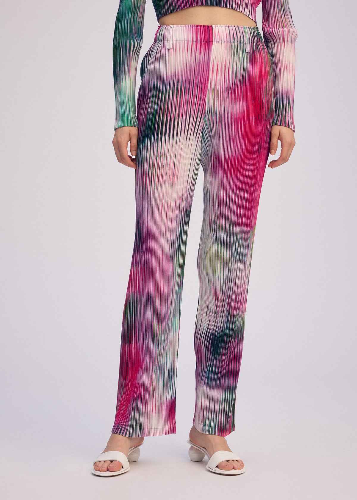 A person wears ISSEY MIYAKE's OBSCURE PLEATS: a colorful moiré-style pleated polyester set in pink, green, and white, paired with white open-toe sandals, standing against a plain light background. view 8 of 8