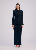 A woman with straight brown hair faces forward against a plain background, wearing the ISSEY MIYAKE HATCHING PLEATS black long-sleeve button-up top and matching pleated straight pants in recycled polyester, paired with black sandals.